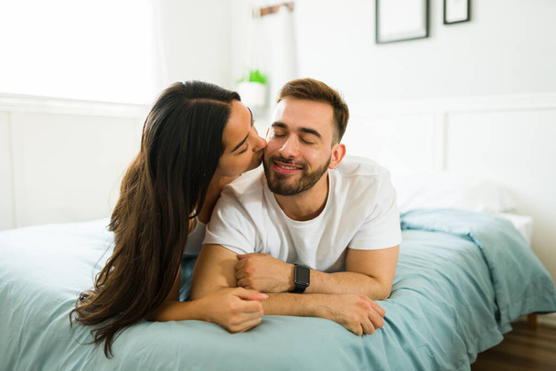 Ihana nuori nainen antaa suukon poskelle onnelliselle aviomiehelleen tai poikaystävälleen nukkuessaan sängyssä.  - Valokuva, kuva