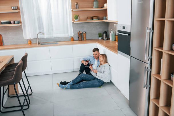 Drinking wine. Couple sitting on the floor of modern kitchen. - Fotografie, Obrázek