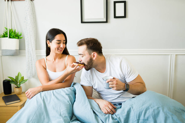 Happy young woman feeding a donut to his partner husband while having breakfast in bed  - Photo, Image