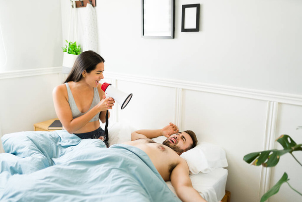 Jovem irritada usando um megafone e gritando para seu marido dormindo na cama pela manhã - Foto, Imagem