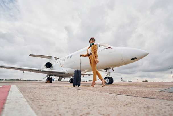 Elegant clothes. Young woman with luggage is outdoors near airplane. - Foto, Imagem