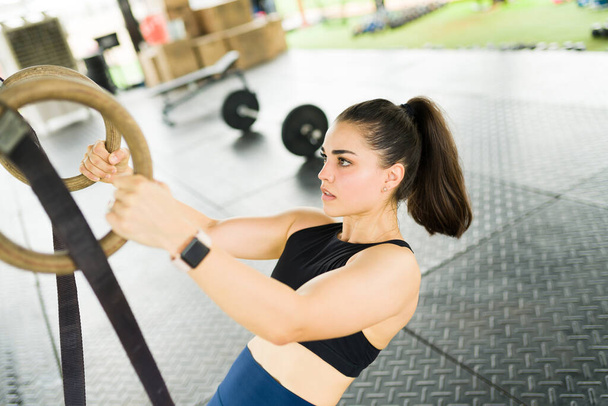 Спортивная женщина выглядит целеустремленной во время использования колец во время тренировки по троеборью и выполнения упражнений на спине - Фото, изображение