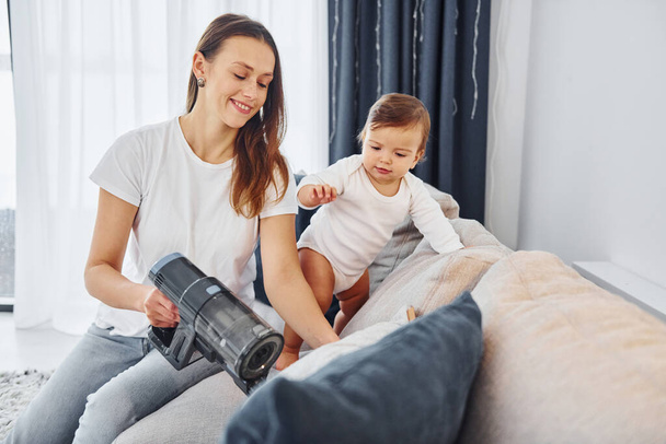 Little part of the vacuum cleaner. Mother with her little daughter is indoors at home together. - Zdjęcie, obraz
