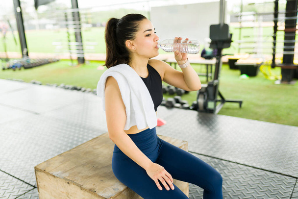 Уставшая молодая женщина, используя полотенце для пота в тренажерном зале и питьевой воды после завершения тренировки упражнения  - Фото, изображение