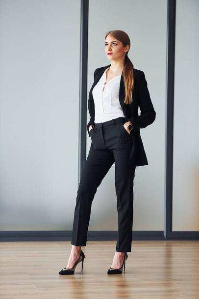 Standing against grey background. Young adult woman in formal clothes is indoors in the office. - Φωτογραφία, εικόνα