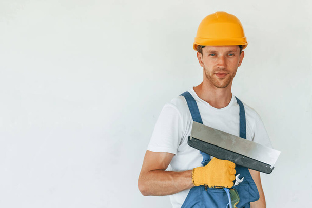 Standing near white wall. Young man working in uniform at construction at daytime. - Foto, afbeelding