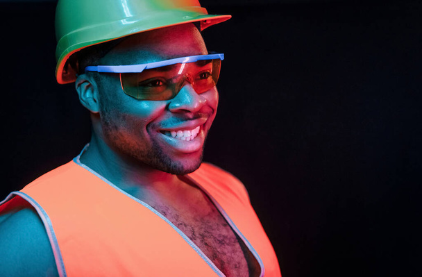 Construction worker in uniform and hard hat. Futuristic neon lighting. Young african american man in the studio. - Φωτογραφία, εικόνα