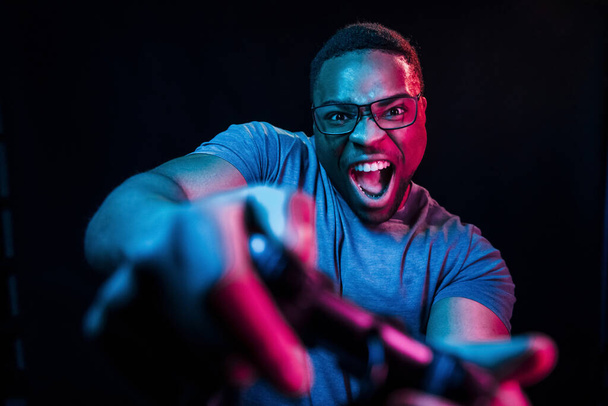 Plays game by using controller. Futuristic neon lighting. Young african american man in the studio. - Photo, image