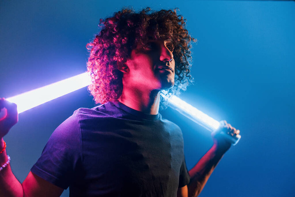 Holds lighting equipment. Young beautiful man with curly hair is indoors in the studio with neon lighting. - Fotografie, Obrázek