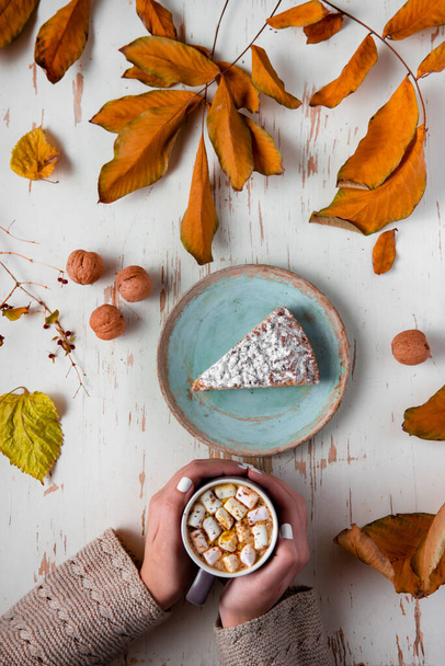 Top view of female hands holding marshmallows on shabby table with piece of cake, walnuts and scattered leaves - Foto, imagen