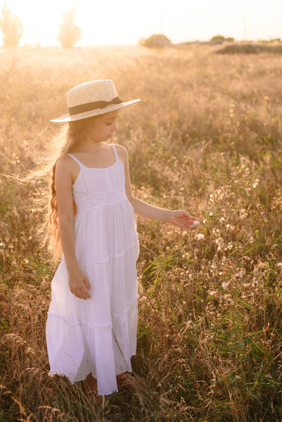 Cute little girl with blond long hair in a summer field at sunset with a white dress with a straw hat - Фото, зображення