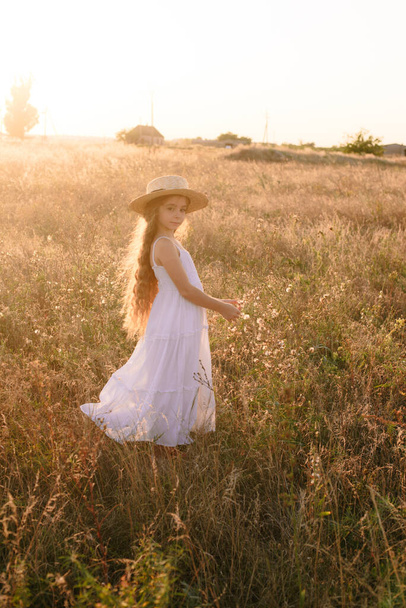 Roztomilá holčička se světlými dlouhými vlasy v letním poli při západu slunce s bílými šaty s slamákem - Fotografie, Obrázek