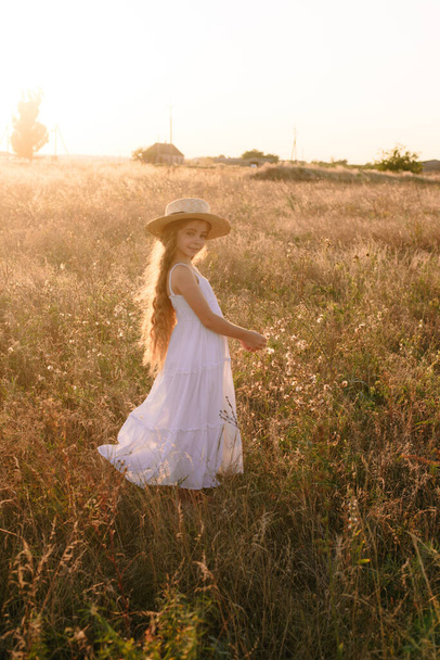 Χαριτωμένο κοριτσάκι με ξανθά μακριά μαλλιά σε ένα καλοκαιρινό χωράφι στο ηλιοβασίλεμα με ένα λευκό φόρεμα με ψάθινο καπέλο - Φωτογραφία, εικόνα