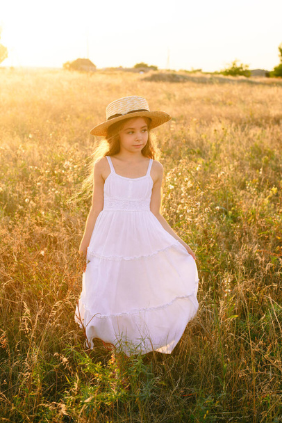Cute little girl with blond long hair in a summer field at sunset with a white dress with a straw hat - Foto, Bild