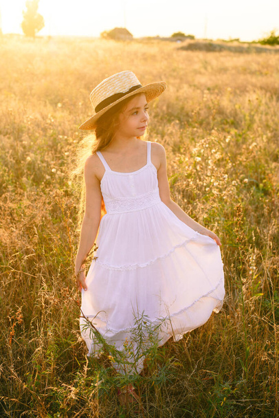 Cute little girl with blond long hair in a summer field at sunset with a white dress with a straw hat - Foto, immagini