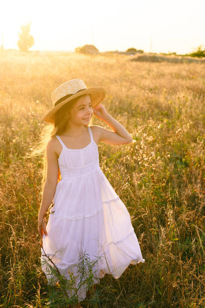 Cute little girl with blond long hair in a summer field at sunset with a white dress with a straw hat - Valokuva, kuva