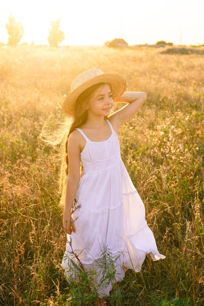 Cute little girl with blond long hair in a summer field at sunset with a white dress with a straw hat - Photo, image