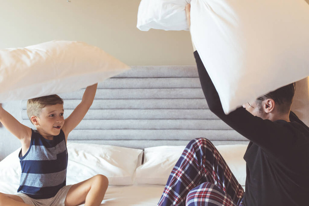 Zabawny chłopiec i jego tata dobrze się bawią podczas walki na poduszki w sypialni. - Zdjęcie, obraz