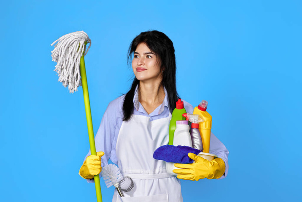 pretty woman in yellow rubber gloves and cleaner apron holding bucket of detergents and mop on blue background. - Photo, image