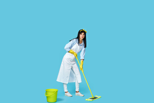 woman in rubber gloves and cleaner apron washing floor with mop on blue background. Full length - Fotoğraf, Görsel