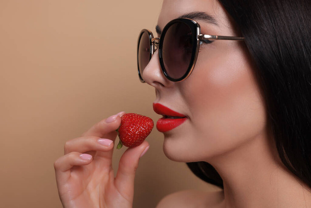 Attractive woman in fashionable sunglasses holding strawberry against beige background, closeup. Space for text - Photo, image