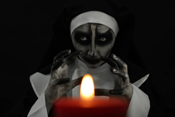 Scary devilish nun with burning candle on black background. Halloween party look - 写真・画像