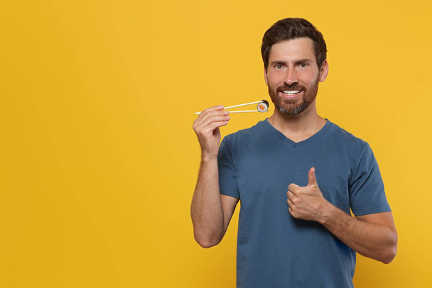 Happy man holding sushi roll with chopsticks and showing thumbs up on orange background. Space for text - Fotografie, Obrázek
