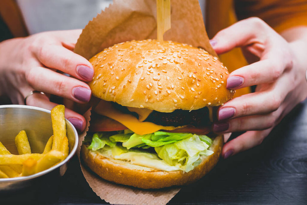 Weibliche Hände halten frische leckere Burger mit Pommes auf dem schwarzen Holztisch - Foto, Bild