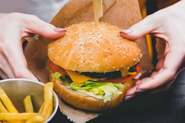 Manos femeninas sosteniendo una deliciosa hamburguesa fresca con papas fritas en la mesa de madera negra - Foto, Imagen