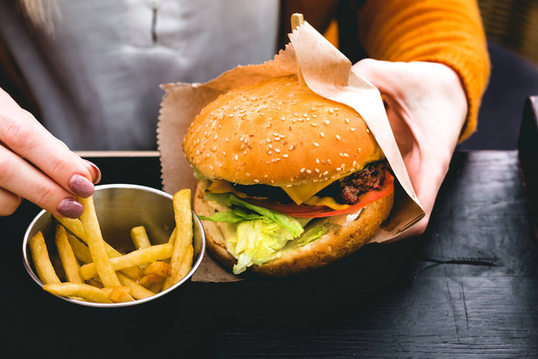 Manos femeninas sosteniendo una deliciosa hamburguesa fresca con papas fritas en la mesa de madera negra - Foto, Imagen