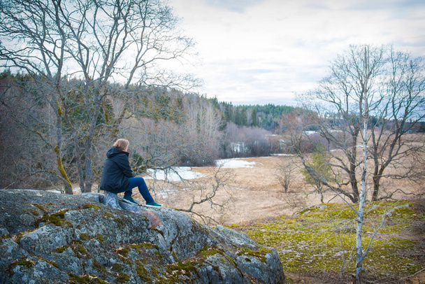 Tavaszi délután, egy lány kaszával ül egy szikla szélén, kék farmerben és fekete kabátban, oldalnézetből.. - Fotó, kép