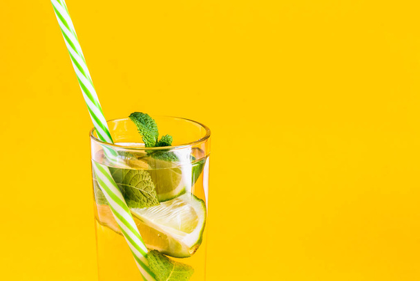Fresh summer lemonade with water, lime and mint in a glass tumbler with a straw on a yellow background. Cold, refreshing summer cocktail close up. Fresh drink with copy space - Foto, imagen