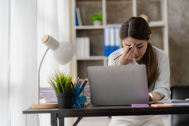 Азиатская бизнесвумен страдает головной болью во время работы над ноутбуком, работающим в офисе финансовой отчетности - Фото, изображение
