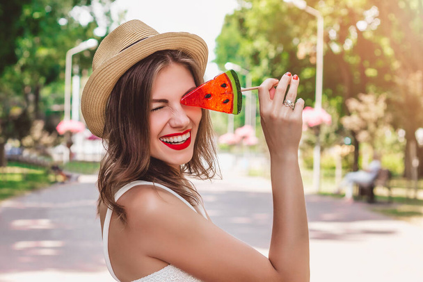 A young girl walks in the park with a lollipop in the form of watermelon. Girl in straw hat smiling in the park - Photo, Image