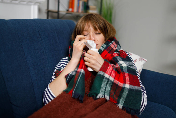 Mujer enferma con una nariz mocosa acostada en un sofá debajo de una manta con un pañuelo - Foto, imagen