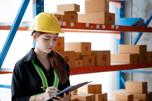 Asian female worker wearing safety clothing is in a warehouse, she is checking the goods in the warehouse before shipping. The concept of working in the warehouse and working in the warehouse is safe. - Photo, Image