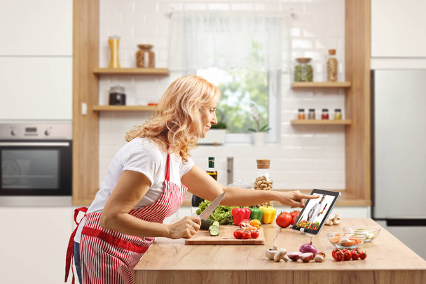 Mature woman cooking on a counter and watching a video on a digital tablet inside a kitchen - Foto, Imagem