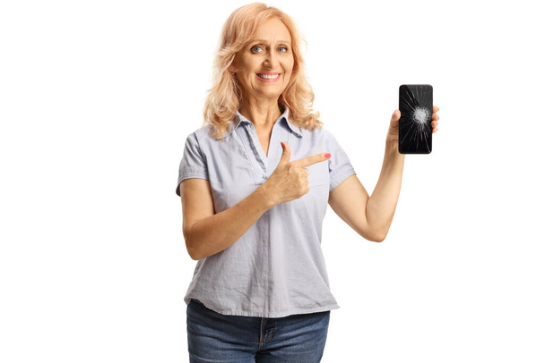 Casual mature woman holding a smartphone and pointing at the broken screen isolated on white background - Photo, Image