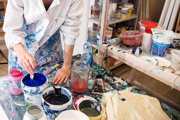 Cropped photo of hands of woman in white robe covered with various stains stirring blue acrylic paint in bucket with brush on desk in workshop. Modern art, creativity, decoration, workshop, painting. - Photo, Image