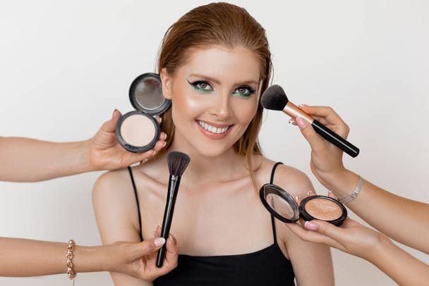 Photo closeup of pretty smiling girl with evening makeup and highlighting cheekbones on white background. Makeup artists applying makeup with brushes and face powder. Professional beauty products. - Foto, Bild