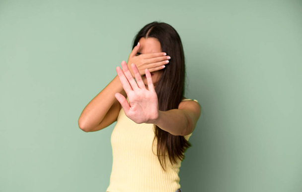 hispánská hezká žena zakrývající obličej rukou a dávající druhou ruku dopředu, aby zastavila kameru, odmítající fotografie nebo obrázky - Fotografie, Obrázek