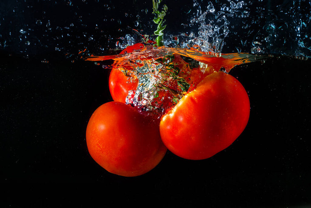 Verse rode tomaat die in water valt met waterspatten en luchtbellen geïsoleerd op zwarte achtergrond. - Foto, afbeelding