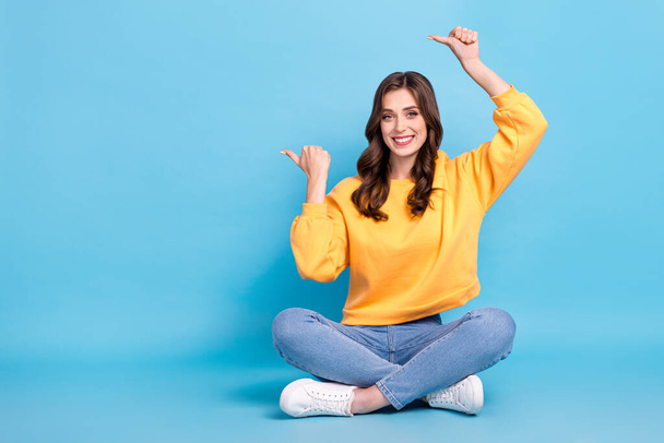 Full length photo of adorable sweet lady wear yellow sweater pointing empty space sitting legs crossed isolated blue color background. - Photo, Image