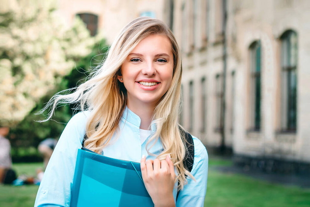 A blonde student girl is smiling and holding a folder and a notebook in her hands at university background. A girl stands on the background of an ancient university, copy space - Фото, изображение
