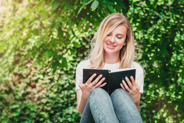 Молода блондинка сидить на лавці, читає книгу і посміхається в парку на фоні дерев і кущів
 - Фото, зображення