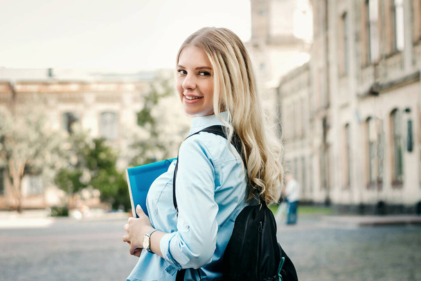 Una studentessa bionda sorride e tiene in mano una cartella e un quaderno su uno sfondo universitario. Ragazza sta prendendo esami all'università - Foto, immagini