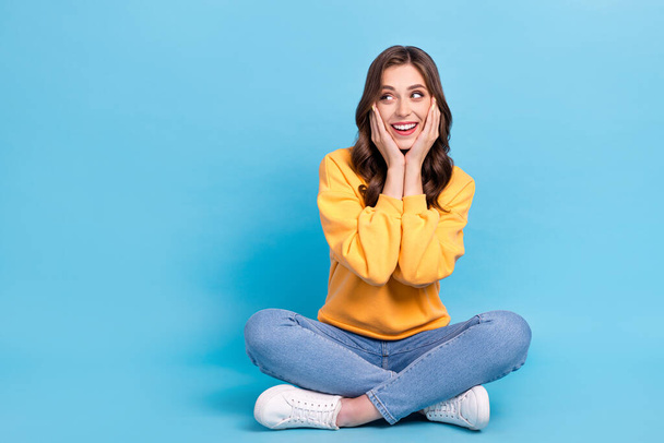 Full length photo of cute dreamy girl dressed yellow pullover sitting legs crossed arms cheeks empty space isolated blue color background. - Photo, Image