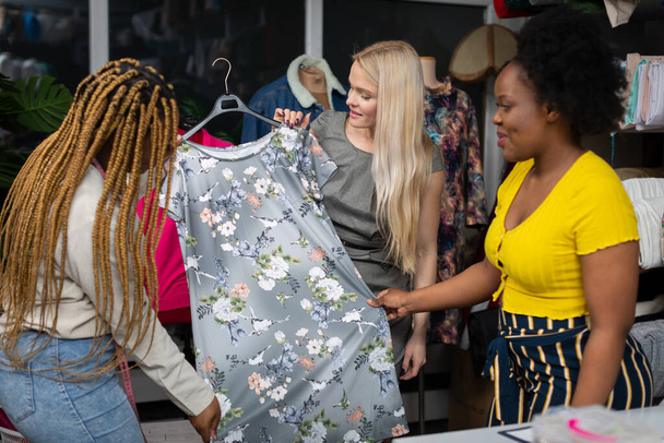 A dark-skinned expeditor shows a customer a new model of dress with a floral pattern. A blonde woman picks out a dress for summer at a tailors shop. - Photo, Image