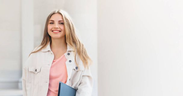 Young blonde student girl smiling against university. Cute girl student holds folders and notebooks in hands, copy space. web banner. copy space - Fotografie, Obrázek