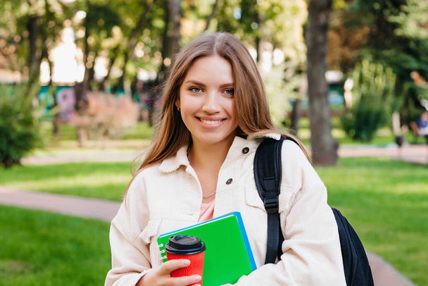 Ξανθιά φοιτήτρια περπατά στο πάρκο με ένα σημειωματάριο και ένα φλιτζάνι καφέ, αντίγραφο χώρο. Εκπαιδευτική έννοια - Φωτογραφία, εικόνα
